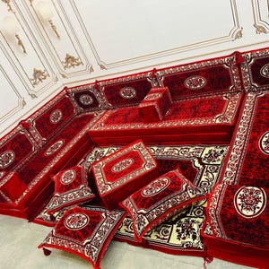 May include: A traditional seating set in red and cream with intricate floral patterns. The set includes a large sofa, several smaller cushions, and a small table. The seating is arranged in a traditional style, with the sofa in the centre and the cushions and table around it.