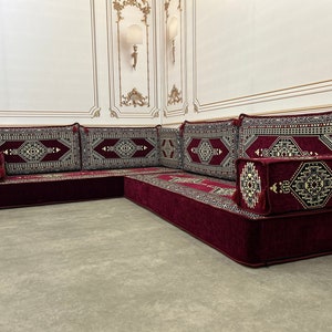 May include: A burgundy-colored L-shaped floor seating set with intricate patterned cushions. The cushions have a geometric design with gold accents. The set is made of a soft, plush fabric.