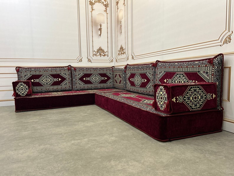 May include: A burgundy-colored L-shaped floor seating set with intricate patterned cushions. The cushions have a geometric design with gold accents. The set is made of a soft, plush fabric.