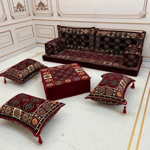 K&ouml;nnte beinhalten: Ein rotes und golden gemustertes Sitzset f&uuml;r den Boden mit einem gro&szlig;en Sofa, einem quadratischen Ottoman und vier Kissen. Das Set ist auf einem wei&szlig;en Boden mit goldenen Verzierungen angeordnet.