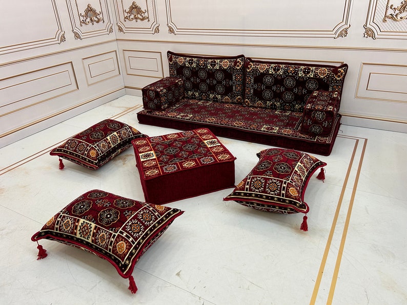 K&ouml;nnte beinhalten: Ein rotes und golden gemustertes Sitzset f&uuml;r den Boden mit einem gro&szlig;en Sofa, einem quadratischen Ottoman und vier Kissen. Das Set ist auf einem wei&szlig;en Boden mit goldenen Verzierungen angeordnet.