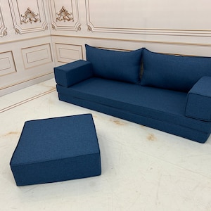 Op de afbeelding: Een donkerblauwe slaapbank met twee kussens en een bijpassende donkerblauwe poef. De slaapbank staat tegen een witte muur met gouden lijstwerk.