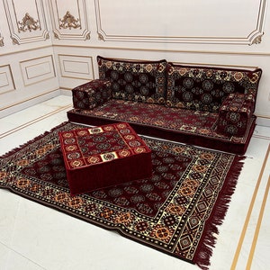 K&ouml;nnte beinhalten: Ein burgunderfarbener Teppich mit goldenen Mustern, ein passender burgunderfarbener Hocker mit goldenen Mustern und ein burgunderfarbenes Sofa mit goldenen Mustern. Das Sofa hat drei Kissen und ist im traditionellen marokkanischen Stil eingerichtet.