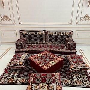 K&ouml;nnte beinhalten: Ein rotes und goldenes Bodenkissen-Set mit passendem Hocker. Das Set beinhaltet ein gro&szlig;es Sofakissen, vier kleinere Kissen und einen quadratischen Hocker. Die Kissen und der Hocker sind alle mit einem pl&uuml;schigen Stoff mit komplizierten geometrischen Mustern bezogen.