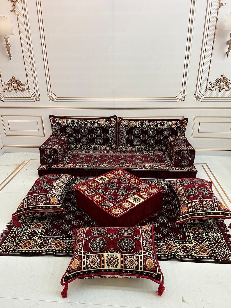 K&ouml;nnte beinhalten: Ein rotes und goldenes Bodenkissen-Set mit passendem Hocker. Das Set beinhaltet ein gro&szlig;es Sofakissen, vier kleinere Kissen und einen quadratischen Hocker. Die Kissen und der Hocker sind alle mit einem pl&uuml;schigen Stoff mit komplizierten geometrischen Mustern bezogen.