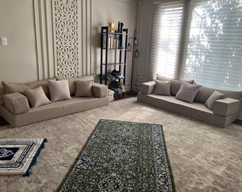 May include: Two beige floor cushions with pillows, arranged as a sofa, in a room with a green and white patterned rug, a bookshelf, and a window with white blinds.