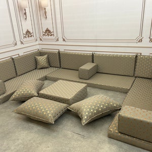 May include: A large, beige sectional sofa with gold patterned upholstery. The sofa is made up of multiple cushions and includes a matching ottoman. The sofa is arranged in a U-shape and is perfect for a living room or family room.