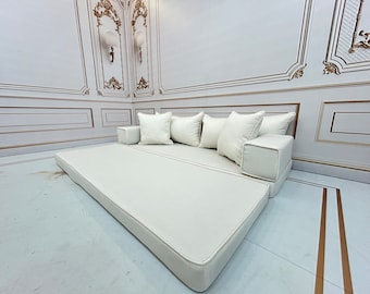 May include: A white, cushioned daybed with a white mattress and four white pillows. The daybed is in a room with white walls and gold trim.