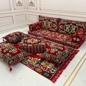 May include: A red and green patterned floor cushion set with a matching sofa. The set includes a large sofa, four square cushions, and a rectangular rug. The cushions and rug have intricate geometric patterns in shades of red, green, and cream.