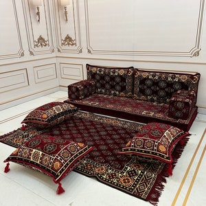 K&ouml;nnte beinhalten: Ein rotes und goldenes Bodenkissen-Set mit einem gro&szlig;en Sofa und vier kleineren Kissen. Die Kissen sind alle aus einem weichen, pl&uuml;schigen Stoff gefertigt und haben komplizierte Muster.