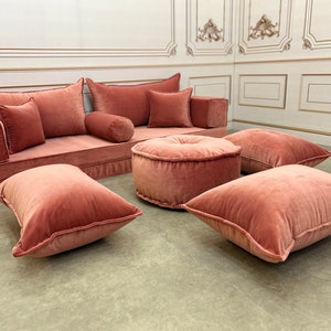 May include: A pink velvet sofa with multiple pillows and two round pink velvet ottomans. The sofa and ottomans are arranged in a living room setting.