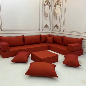 May include: A red corner sofa with a matching ottoman and throw cushions. The sofa is made of a soft, plush fabric and is ideal for relaxing and entertaining guests.