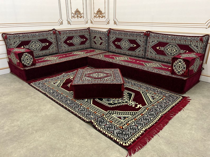 May include: A burgundy red sectional sofa with intricate patterned cushions and a matching burgundy red rug with a geometric pattern. The sofa is set up in a corner and includes a small square ottoman.