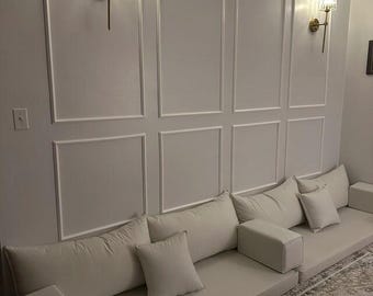 May include: A modern living room setup featuring a light-colored sofa with multiple rectangular and square pillows. The sofa is placed against a white wall with decorative paneling. Two gold sconces provide lighting.