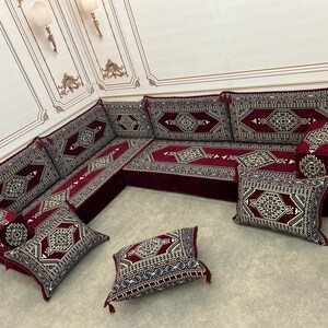 May include: A burgundy and cream colored traditional Moroccan floor seating set with intricate geometric patterns. The set includes a large L-shaped sofa with multiple cushions and several smaller throw pillows.