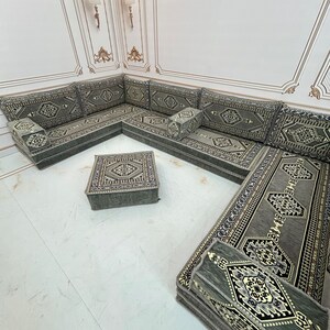 May include: A traditional Moroccan floor seating set with a green and gold patterned fabric. The set includes a large L-shaped sofa, a square ottoman, and several smaller cushions.