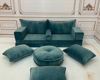 May include: A teal blue velvet sofa with four matching cushions and a round ottoman. The sofa has a low back and armrests, and the cushions are all square except for the ottoman, which is round. The sofa is sitting on a white floor.