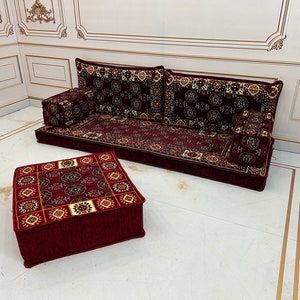 K&ouml;nnte beinhalten: Ein rotes und cremefarbenes Bodenkissen-Set mit einer passenden quadratischen Ottomane. Die Kissen sind in Sofaform mit R&uuml;ckenlehne und zwei Armlehnen angeordnet. Das Set befindet sich auf einem wei&szlig;en Boden mit goldenen Verzierungen.