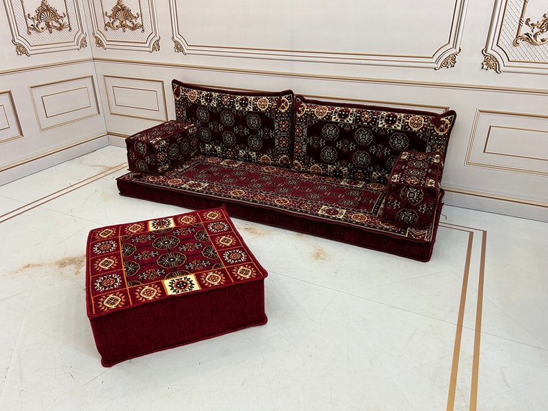 K&ouml;nnte beinhalten: Ein rotes und cremefarbenes Bodenkissen-Set mit einer passenden quadratischen Ottomane. Die Kissen sind in Sofaform mit R&uuml;ckenlehne und zwei Armlehnen angeordnet. Das Set befindet sich auf einem wei&szlig;en Boden mit goldenen Verzierungen.