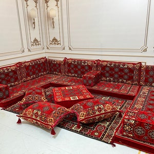 Puede incluir: Un conjunto de asientos de suelo con estampado rojo y crema con múltiples cojines y un pequeño otomano cuadrado. El conjunto está dispuesto en un estilo tradicional con una gran área de asientos en forma de L y un pequeño otomano en el centro.