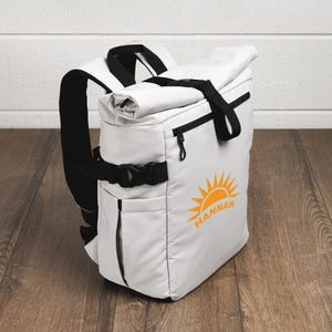 May include: A white roll-top backpack featuring black straps, black zippers, and a side pocket. The front displays an orange graphic of a rising sun with the name HANNAH printed in orange capital letters beneath the sun icon on the bag surface.
