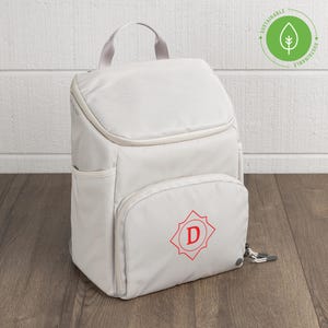 May include: A light gray backpack featuring a top handle, a front zippered pocket with a red letter D inside a star-shaped emblem, and side pockets. A green circular logo with a leaf icon and the word SUSTAINABLE is visible in the upper corner.