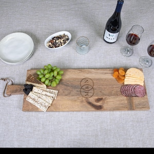 May include: A wooden serving board with a handle, featuring a selection of food items. The board holds grapes, cheese, crackers, salami, and dried apricots. A bottle of wine and two glasses of red wine are also present. The board is engraved with the letters "M & N".