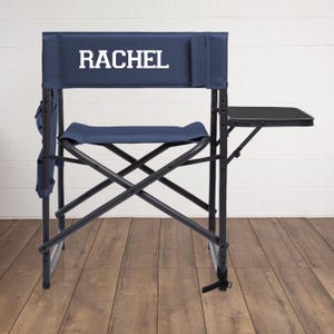May include: A navy blue director's chair with the name "RACHEL" printed in white on the backrest. The chair has a black frame, a navy blue seat and back, and a black side table. The chair is set against a white wood-paneled wall.