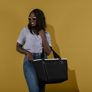 Puede incluir: Una persona con una camisa blanca y pantalones vaqueros azules sostiene una bolsa de mano negra con costuras blancas. La bolsa tiene un forro blanco y un bolsillo grande en la parte delantera.