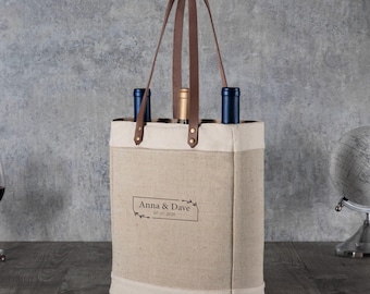 May include: A beige canvas wine tote bag with two brown leather handles and a rectangular border print containing the text Anna & Dave 07-17-2020. The bag holds three wine bottles with blue and gold foil tops visible at the top opening.