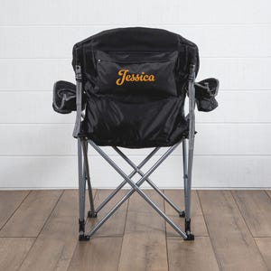 May include: A black folding chair with a gray frame and a black fabric seat. The chair has a pocket on the back with the name "Jessica" embroidered in orange.