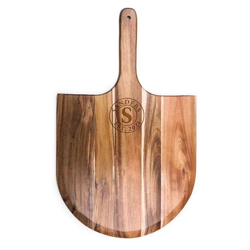 May include: A wooden pizza peel with a long handle and a rounded base. The wood features natural brown grain patterns. Engraved in the center is a circular design containing the letter S, the word SANDERS, and the text EST. 2021.