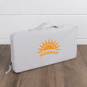 May include: A light gray rectangular bag with a handle and a zipper closure. The bag features an orange sunburst design with the name "HANNAH" printed below it. The bag is sitting on a wooden surface.