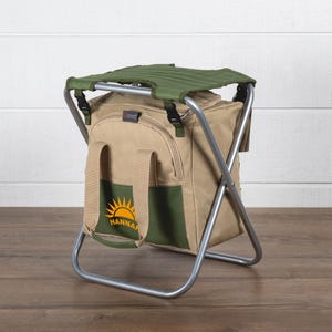 May include: A tan and green portable folding chair with a cooler bag. The chair has a green seat and a silver metal frame. The cooler bag has a green pocket with a yellow sun logo and the name "HANNAH".
