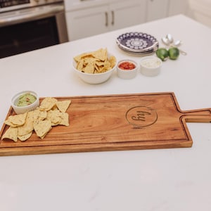 May include: A rectangular wooden serving board with a handle, featuring a laser-engraved circular logo with the letters "hy" inside. The board is topped with a small white bowl of guacamole and tortilla chips, alongside other bowls of snacks.