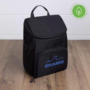 May include: A black backpack with a top handle and a front zippered pocket. The front pocket features a blue graphic of a wave above the name EDUARDO. A green circular sustainable logo is visible in the upper right corner of the image.