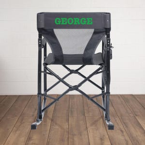 May include: A black folding camping chair with the name "GEORGE" printed in green on the backrest. The chair has a mesh back and seat, a side pocket, and a black metal frame. The chair is designed for outdoor use.