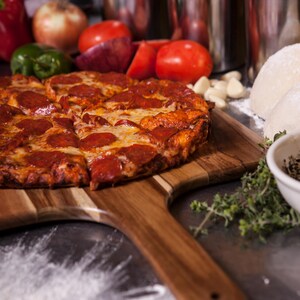May include: A round pepperoni pizza with melted cheese on a wooden serving paddle. The pizza is sliced and features a golden-brown crust. Nearby are fresh red tomatoes, onions, green peppers, garlic cloves, dough balls, and sprigs of fresh herbs.