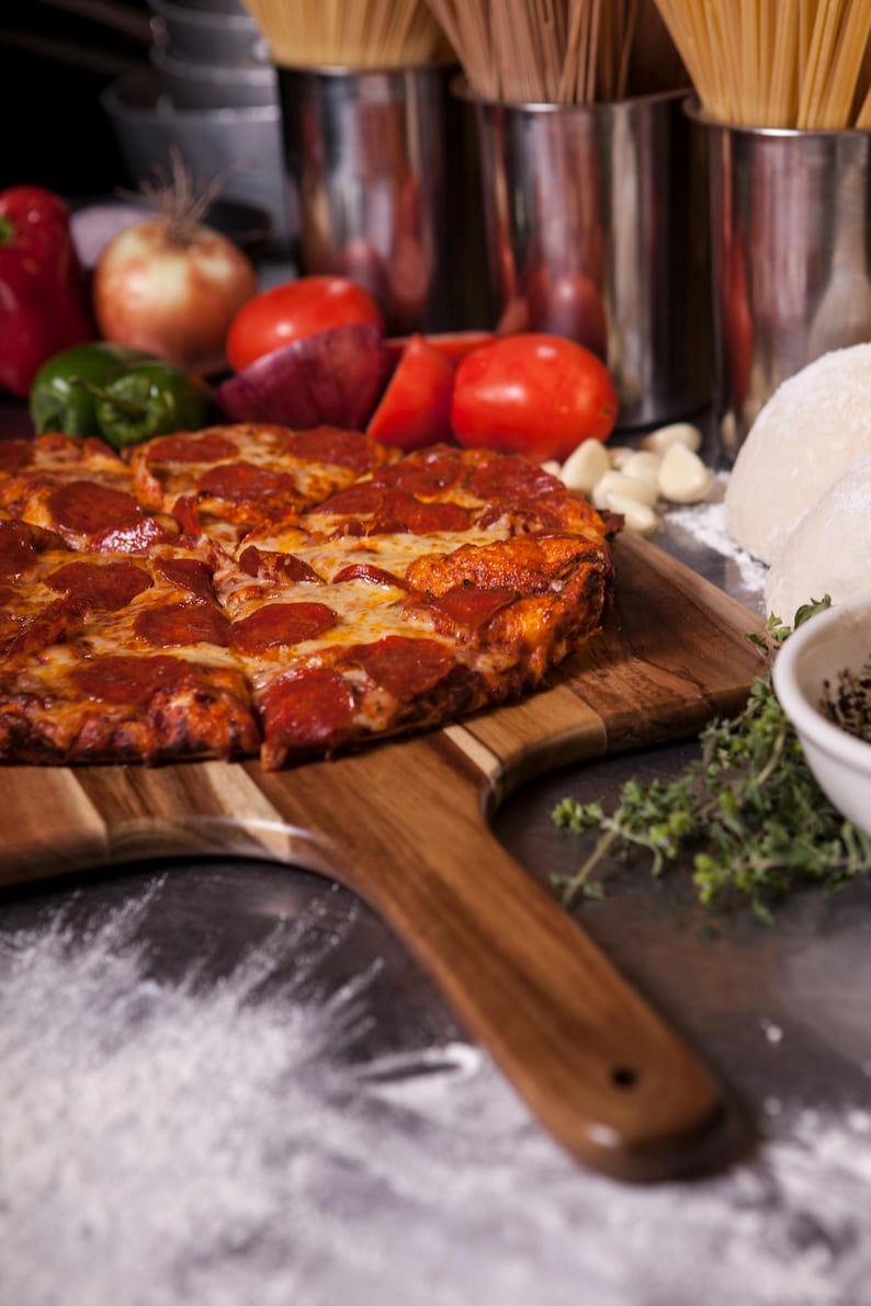 May include: A round pepperoni pizza with melted cheese on a wooden serving paddle. The pizza is sliced and features a golden-brown crust. Nearby are fresh red tomatoes, onions, green peppers, garlic cloves, dough balls, and sprigs of fresh herbs.