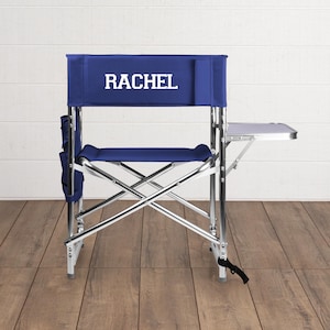 May include: A navy blue director's chair with the name "RACHEL" printed in white on the backrest. The chair has a silver metal frame, a side table, and a side pocket. The chair is designed for outdoor use.