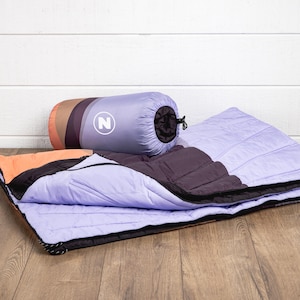 May include: A lavender and black sleeping bag with a matching rolled-up carrying case. The case has a lavender body with a white "N" logo and a striped end in shades of brown and orange. The sleeping bag is open, showing a black interior.