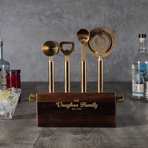 May include: A bar tool set with gold-colored tools and a dark wooden base. The set includes a jigger, bottle opener, spoon, and strainer. The base is engraved with "The Vaughan Family EST 1993".