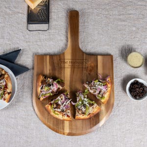 May include: A wooden pizza peel with a handle, engraved with "The Hudson's 09.28.1987". The peel is on a light beige surface and has four slices of pizza on it.