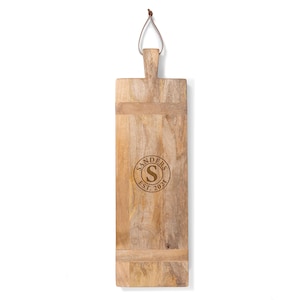 May include: A long, rectangular wooden serving board with a leather loop handle. The board is engraved with the text "SANDERS EST. 2021" and a circle containing the letter "S".