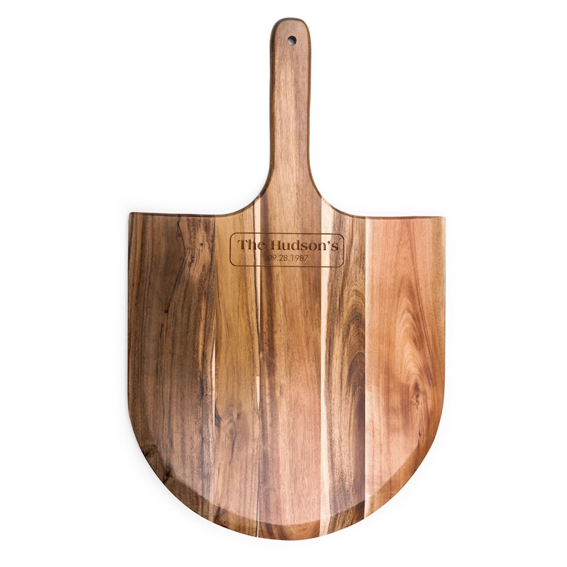 May include: A wooden pizza peel with a long handle and a rounded blade. The wood features natural grain patterns in shades of brown. Engraved in the center is the text The Hudson's 09.28.1987 inside a rectangular border with rounded corners.