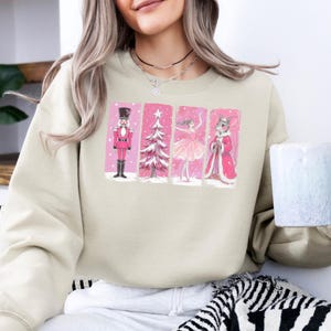 May include: Beige sweatshirt featuring a pink and white graphic of a nutcracker, Christmas tree, ballerina, and mouse king. The illustrations are set against a pink background. The sweatshirt has a crew neck and long sleeves.