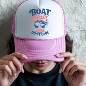 May include: Pink and white trucker hat with the text "BOAT HAIR DON'T CARE" in blue and coral. The hat features a graphic of sunglasses and a boat. The hat has a pink brim and mesh back.