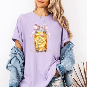 Peut inclure: Un t-shirt violet avec une illustration aquarelle d'un bocal en verre rempli de th&eacute; glac&eacute; et de tranches de citron. Le bocal est d&eacute;cor&eacute; d'un n&oelig;ud rose et d'une paille.