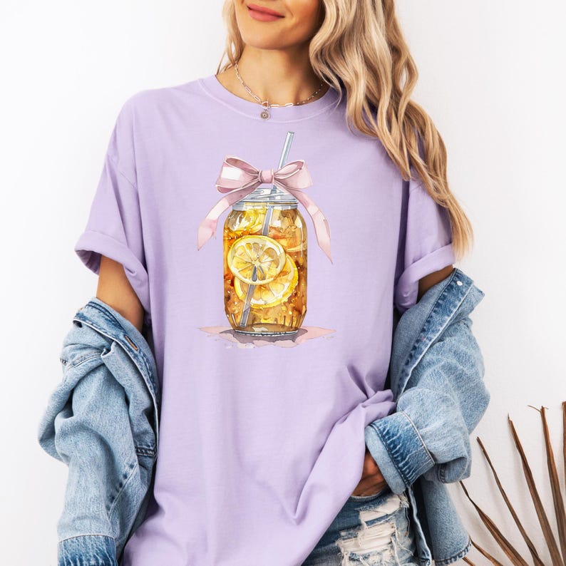 Peut inclure: Un t-shirt violet avec une illustration aquarelle d'un bocal en verre rempli de th&eacute; glac&eacute; et de tranches de citron. Le bocal est d&eacute;cor&eacute; d'un n&oelig;ud rose et d'une paille.