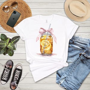 Peut inclure: T-shirt blanc avec une illustration aquarelle d'un bocal en verre rempli de th&eacute; glac&eacute; et de tranches de citron. Un ruban rose est nou&eacute; autour du bocal et une paille sort du haut.